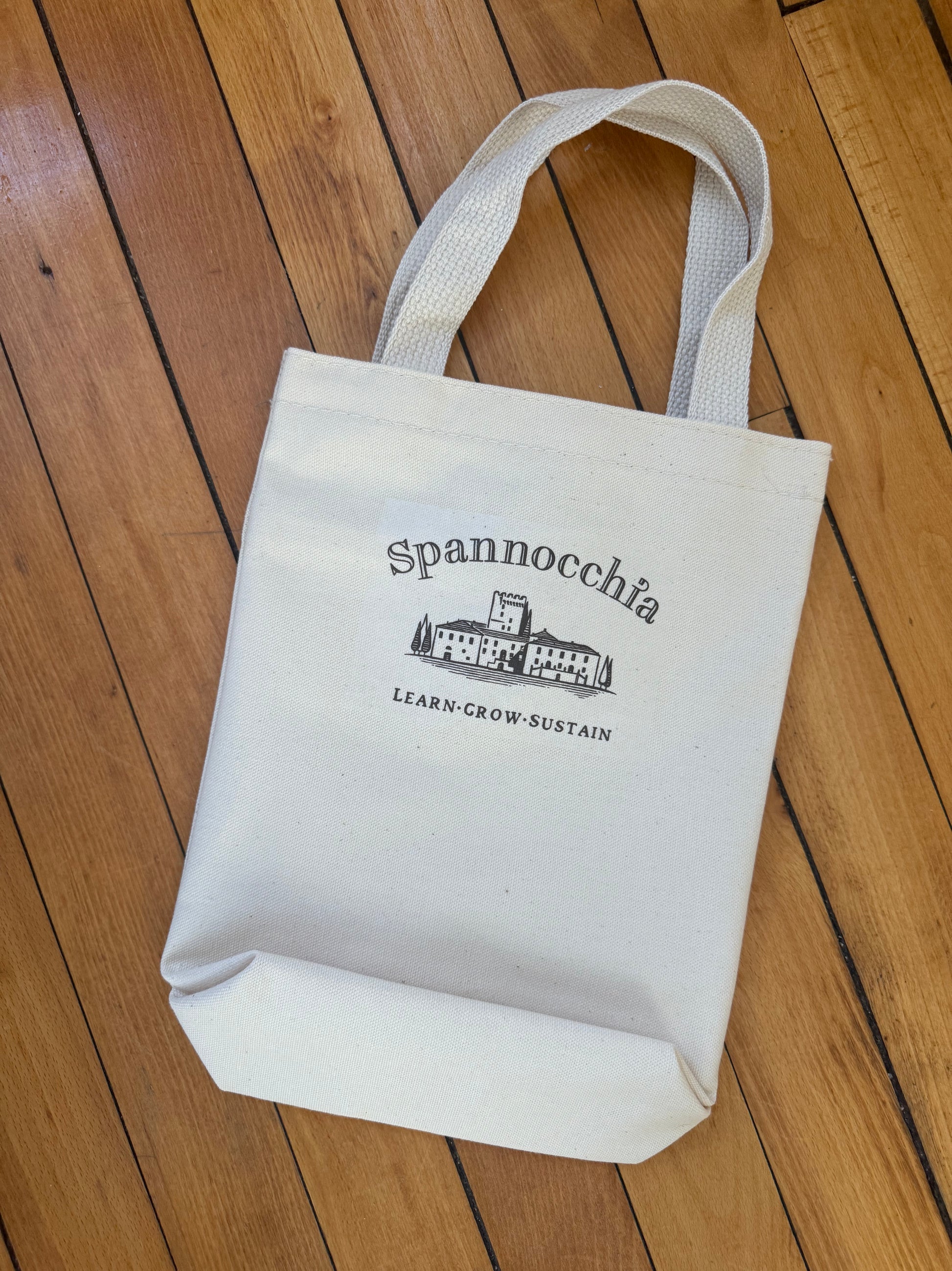 Tote bag showing standard Spannocchia Learn*Grow*Sustain logo against a wood floor.