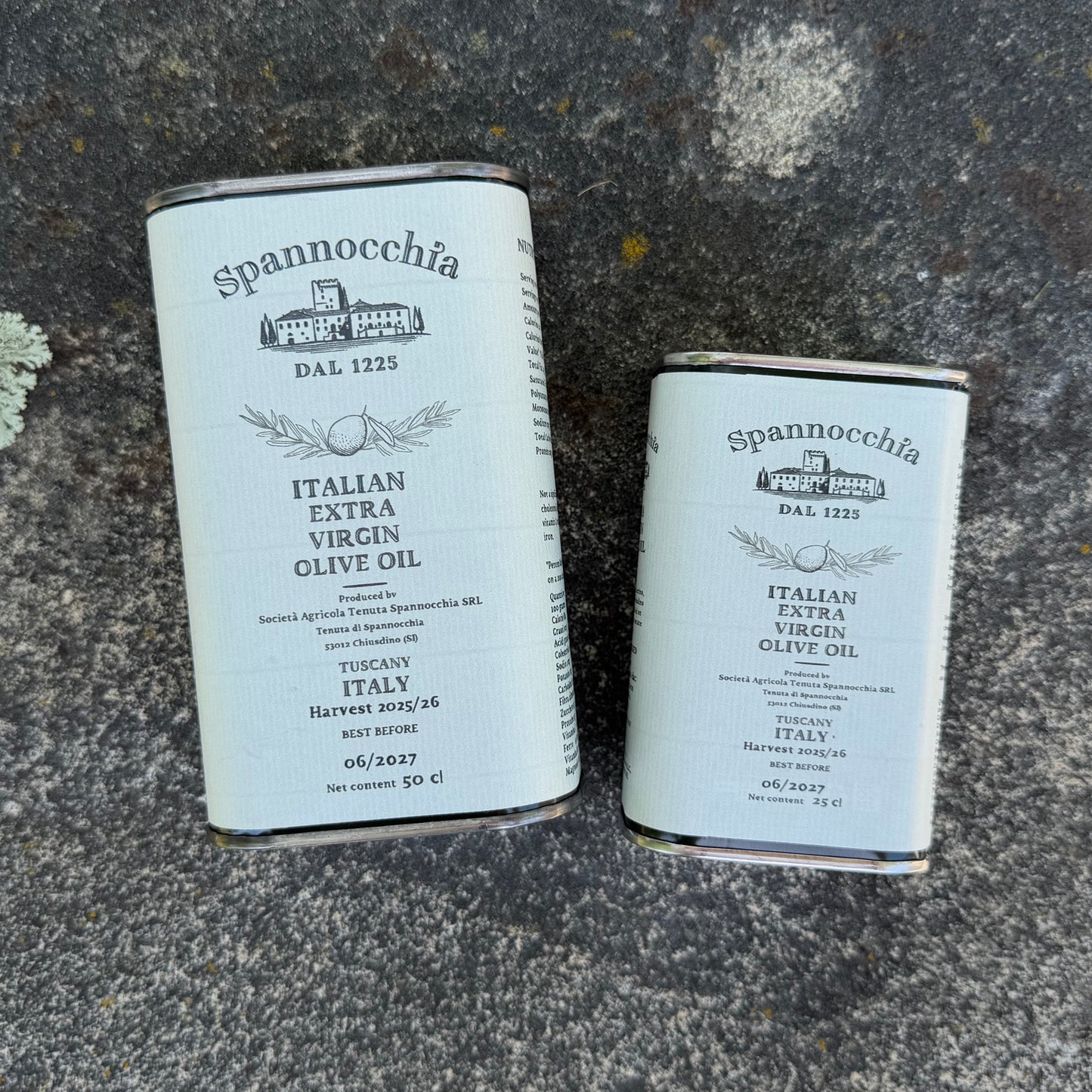 Two cans of Spannocchia olive oil, one large and one small, lying on a rustic stone surface.