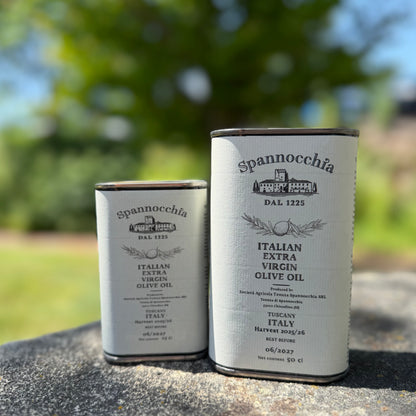 Two cans of Spannocchia olive oil, one large and one small, stand side-by-side on a rustic stone surface with blurred greenery in the background.
