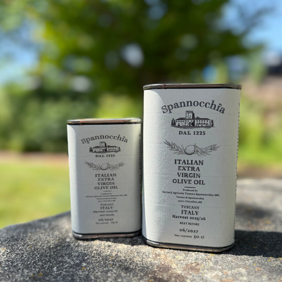 Two cans of Spannocchia olive oil standing side-by-side on a rustic stone surface, with leafy shadows dancing in the breeze and bird song in the background.