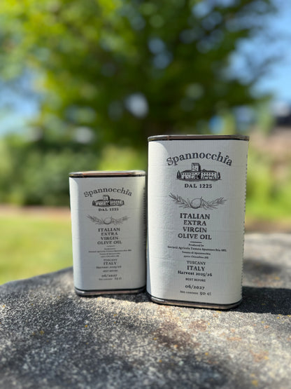 Two cans of Spannocchia olive oil standing side-by-side on a rustic stone surface, with leafy shadows dancing in the breeze and bird song in the background.