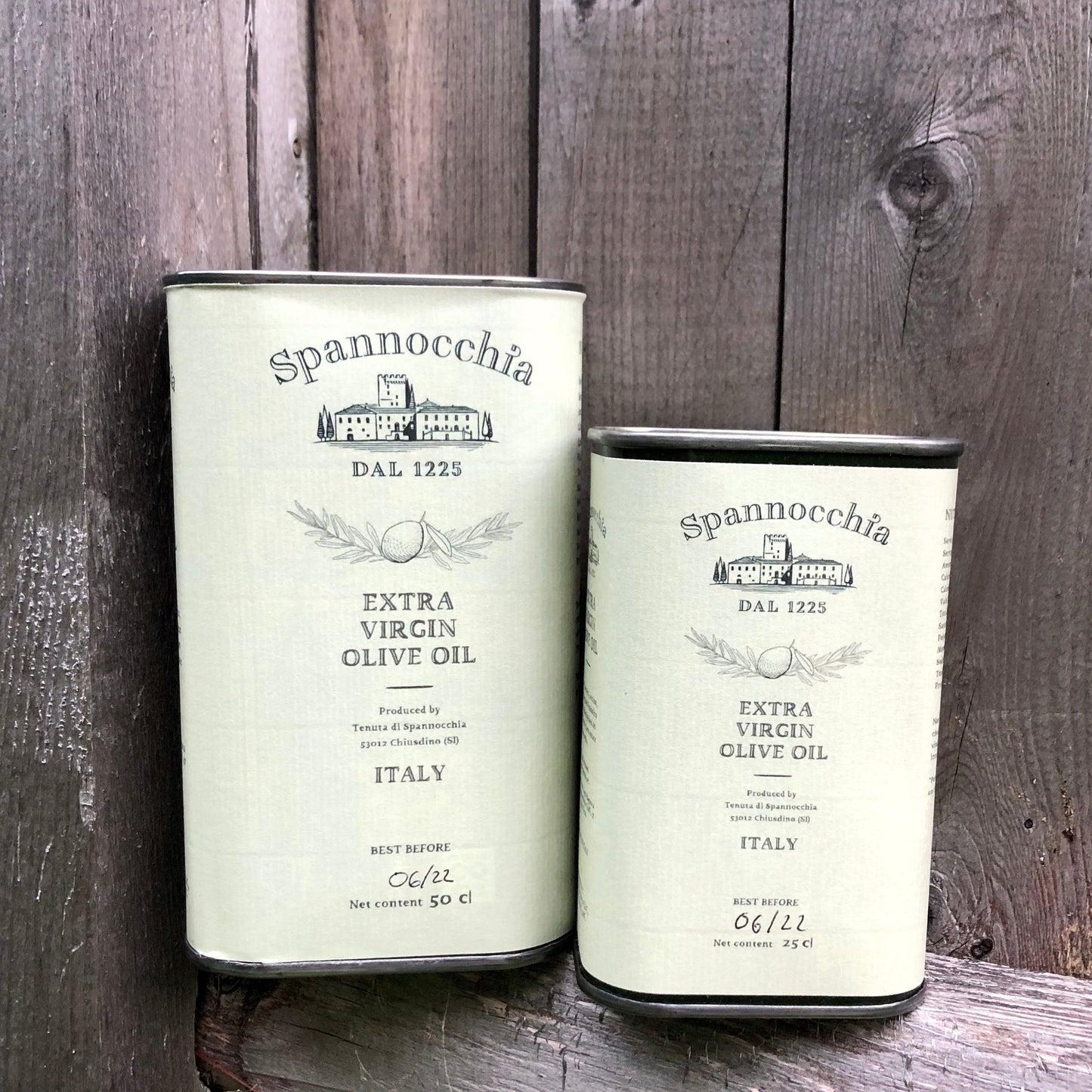 Two cans of Spannocchia olive oil against a weathered wooden fence.